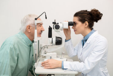 Young brunette female ophthalmologist sitting by medical equipment in clinics while testing eyesight of senior male patient in front of her